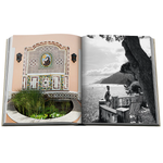An open hardcover book showing two printed photographs on the spread: a left page with a pastel-toned ornate mosaic wall and garden pond, and a right page with a black-and-white coastal scene featuring a person, bench, tree, and water.