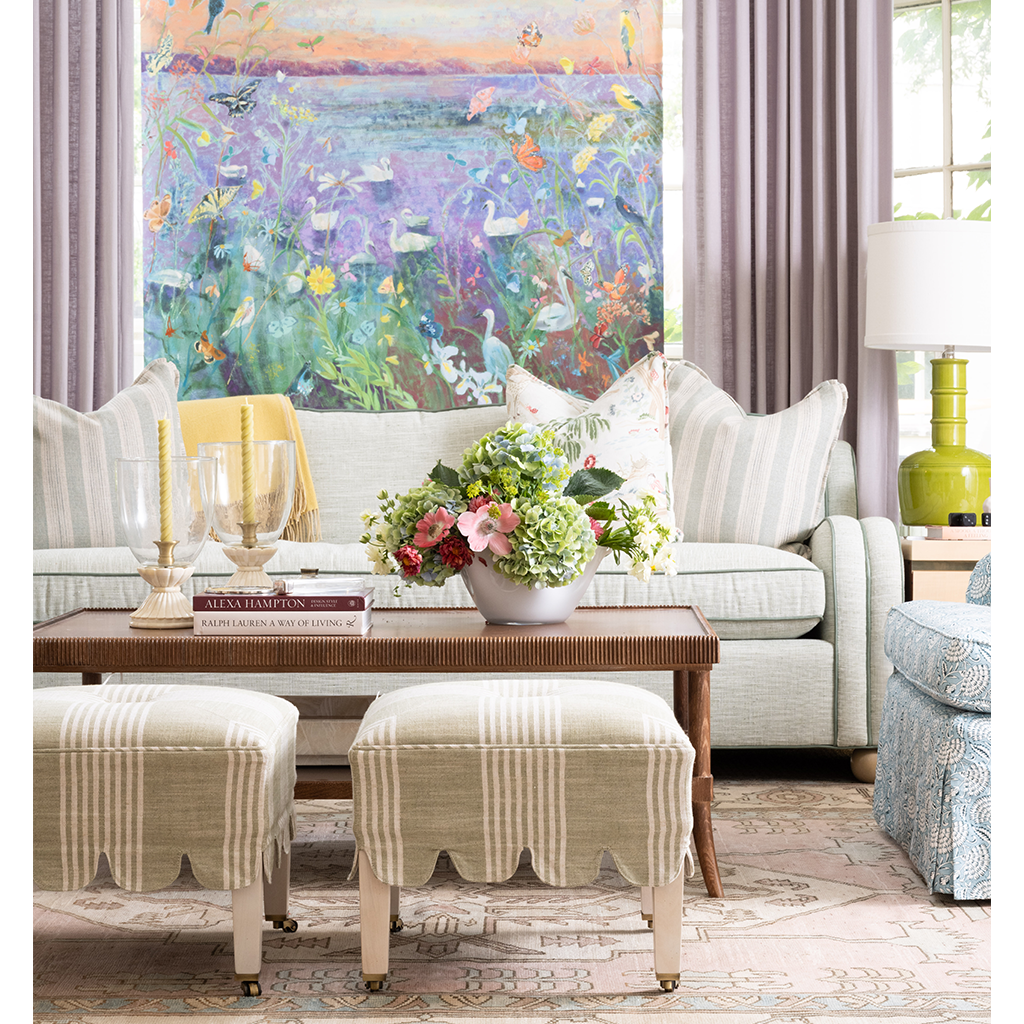 A styled living room featuring a large colorful nature painting on the wall, a light beige sofa with striped pillows, a wooden coffee table, striped ottomans, and a green side table with a lamp.