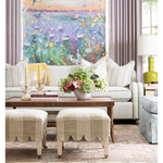 A styled living room featuring a large colorful nature painting on the wall, a light beige sofa with striped pillows, a wooden coffee table, striped ottomans, and a green side table with a lamp.