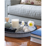 A round gray fabric-covered tray with black braided trim sits on a wooden table in a living room setting, styled with a candle, marble slice, small vase, flowers, and books. A striped sofa is visible in the background.