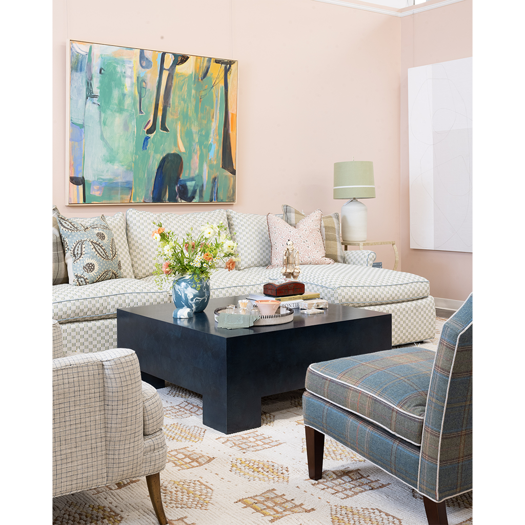 A styled living room scene with a beige sectional sofa, patterned throw pillows, a dark wood coffee table, a blue plaid accent chair, a rug, and a large abstract painting on the wall.