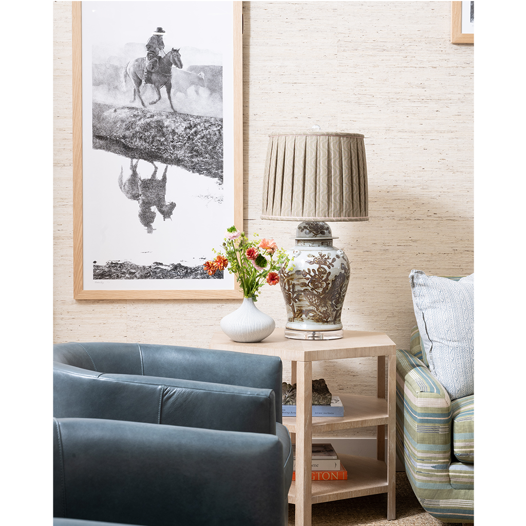 A styled interior corner with a framed black-and-white western landscape print on the wall, a pleated beige table lamp on a light wood side table, a white vase with red flowers, and two upholstered chairs.