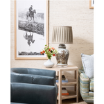 A styled interior corner with a framed black-and-white western landscape print on the wall, a pleated beige table lamp on a light wood side table, a white vase with red flowers, and two upholstered chairs.