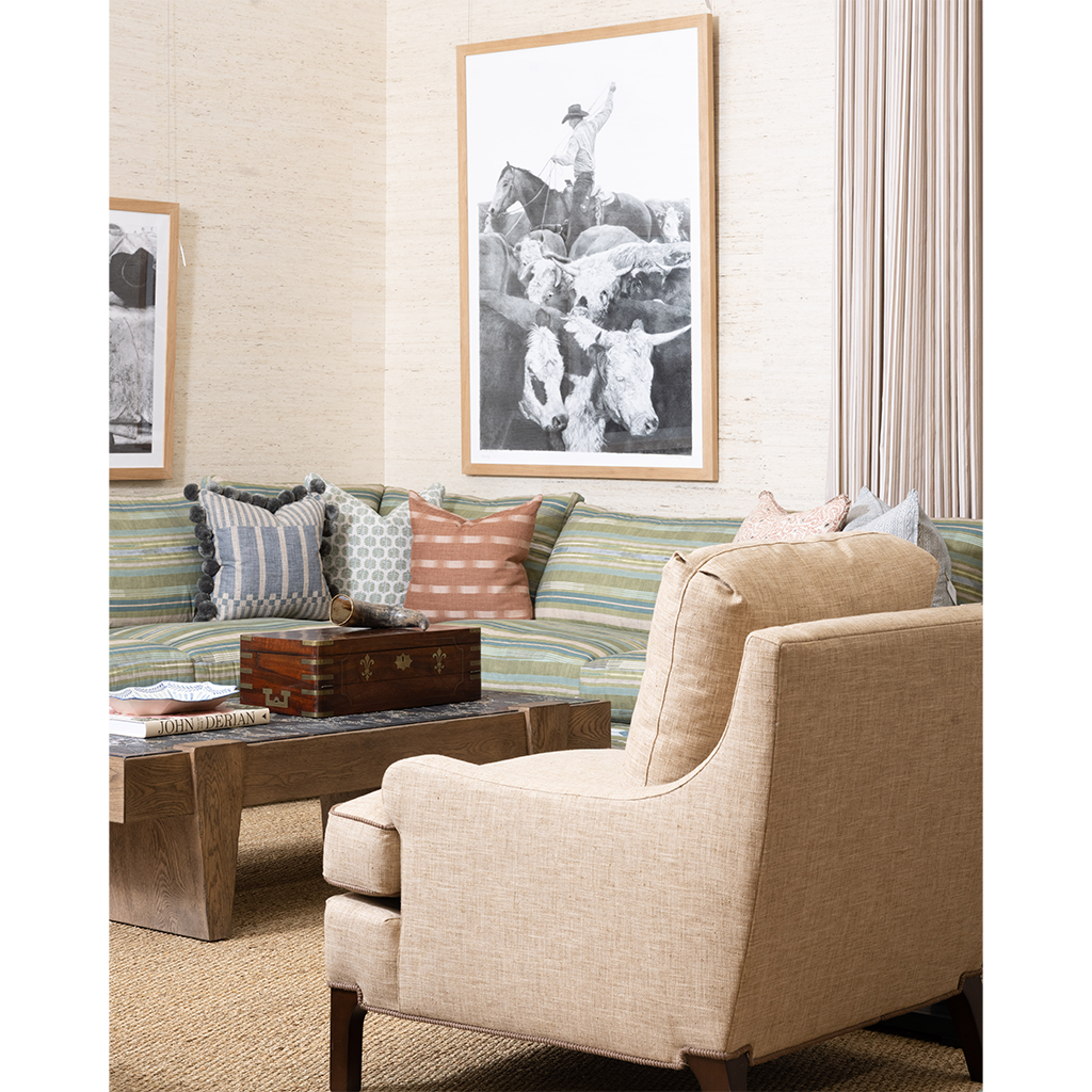 A styled living room scene featuring a beige upholstered armchair in the foreground, a striped green and blue sofa with accent pillows, a wooden coffee table, and framed wall art above the couch.