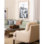 A styled living room scene featuring a beige upholstered armchair in the foreground, a striped green and blue sofa with accent pillows, a wooden coffee table, and framed wall art above the couch.